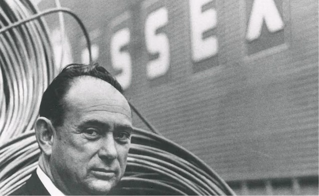 Black and white photo of a man looking at the camera with coiled metal wire and large letters behind him.