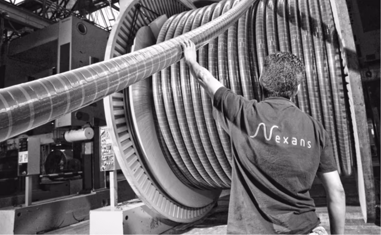 Worker with Nexans shirt handling large industrial cable spool in factory setting.