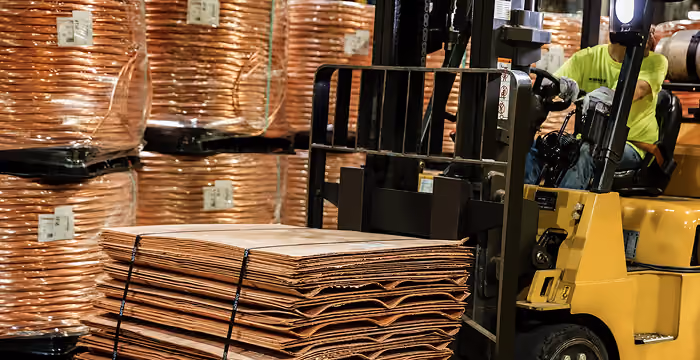Forklift operator moving stacked sheets of copper with large wrapped copper coils in the background.