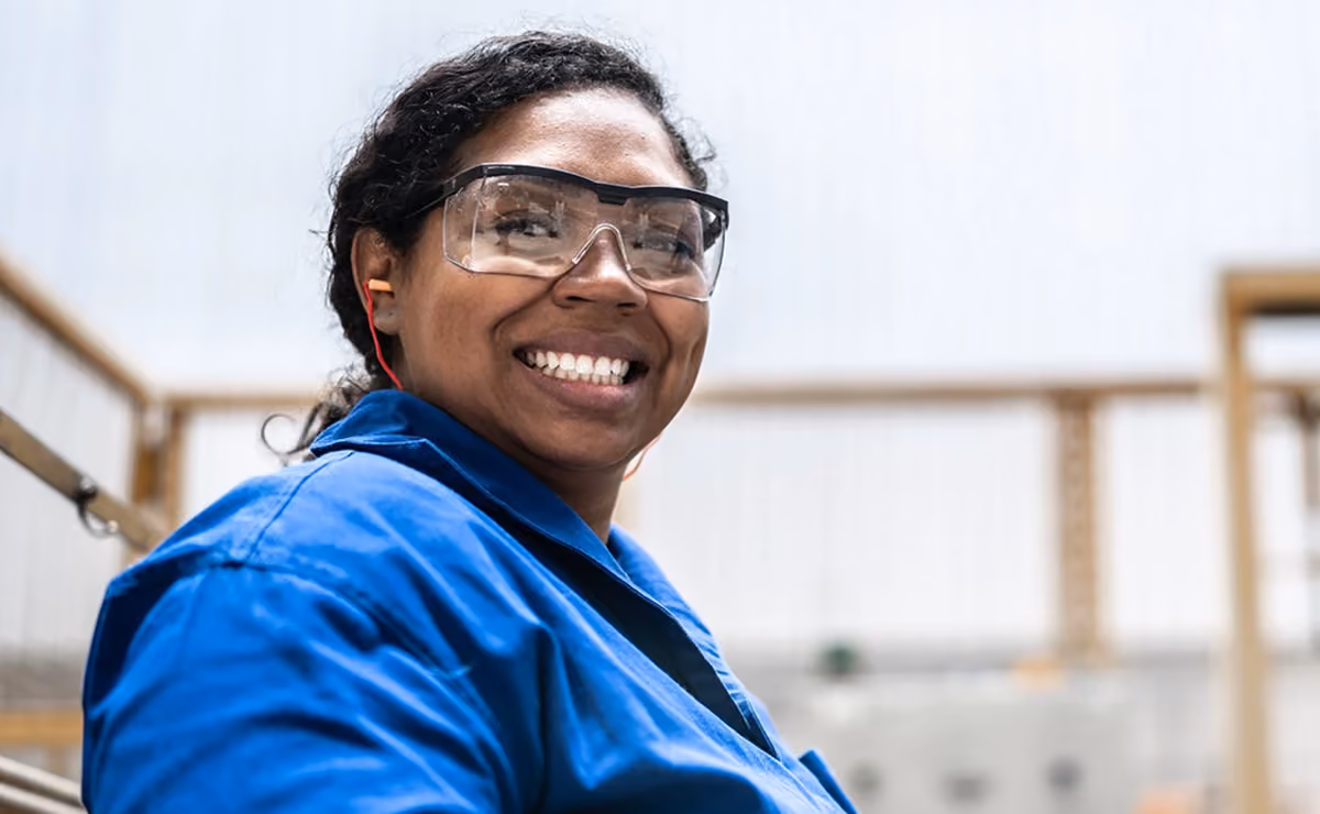 Smiling woman in blue work jacket wearing safety glasses and earplugs in an industrial setting.