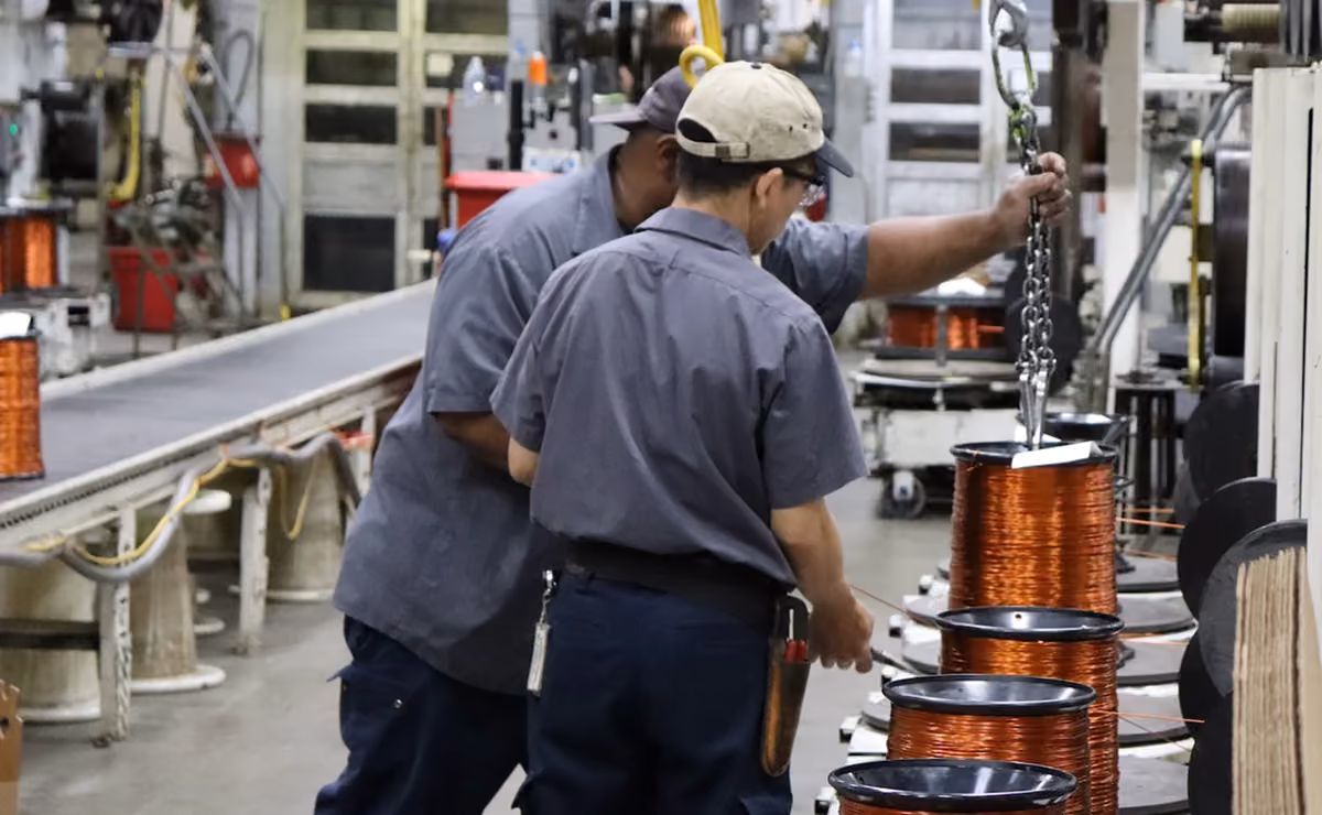 Two factory workers in uniforms operating machinery with large copper wire spools.
