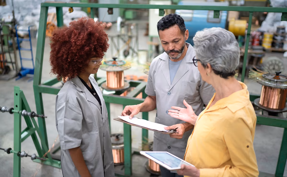Three factory workers discussing paperwork and tablet near industrial equipment with copper coils.