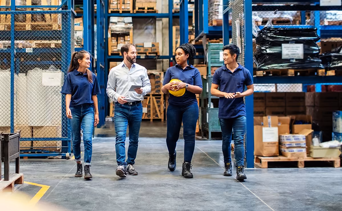 Four warehouse workers walking in aisle, two wearing navy shirts, one holding a yellow helmet.