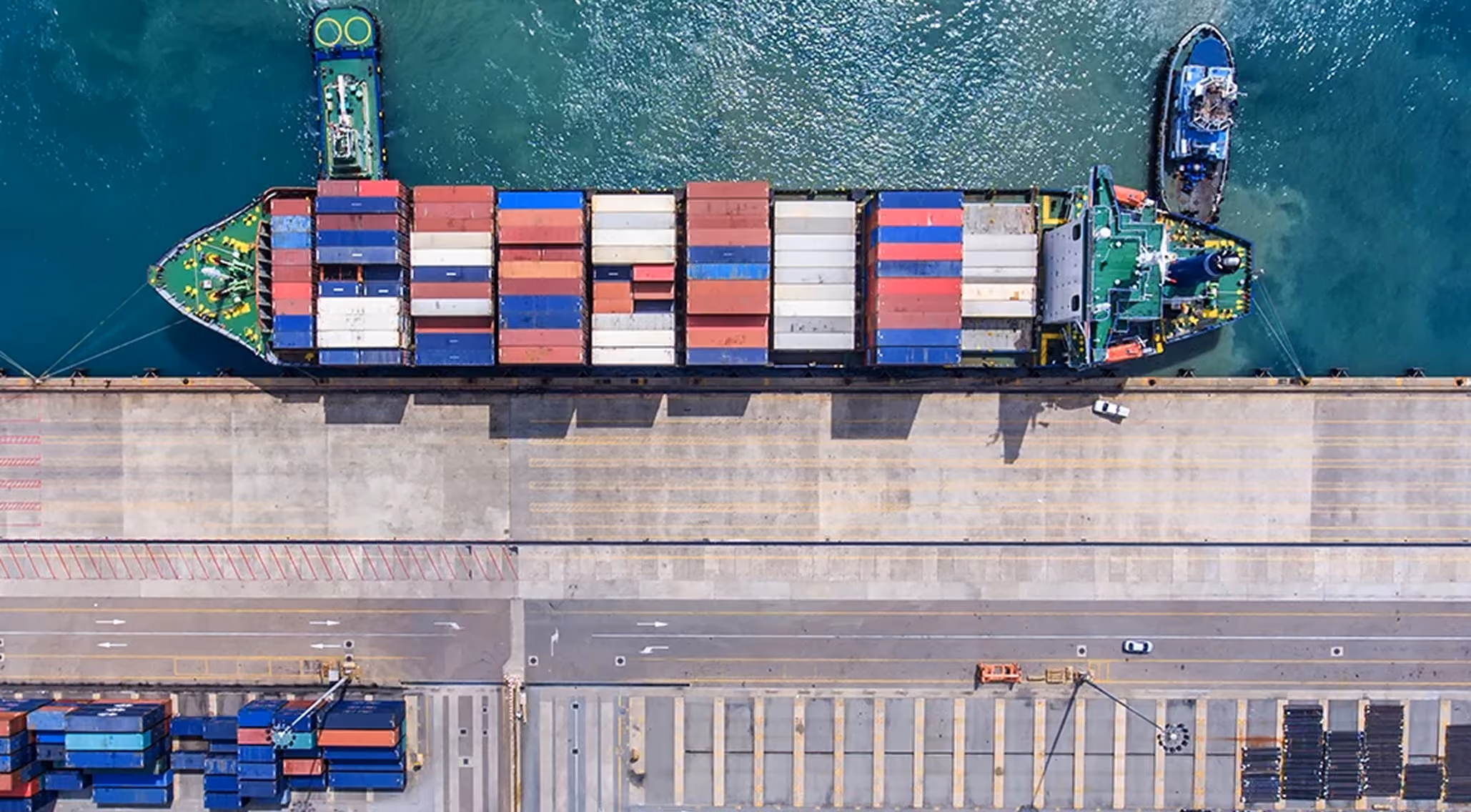 Aerial view of a cargo ship loaded with containers docked at a port with trucks and stacked containers nearby.