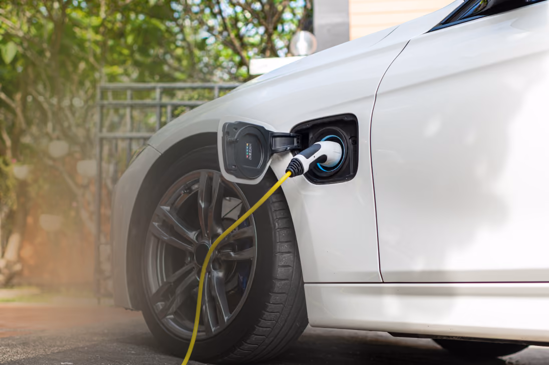 White electric car plugged in and charging with a yellow cable outdoors near trees.