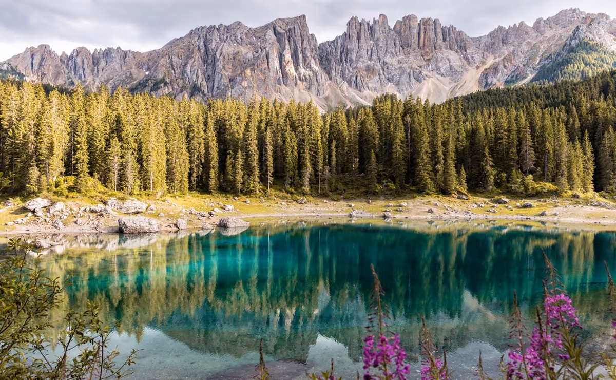 Clear turquoise lake reflecting forest and rocky mountain peaks under cloudy sky.