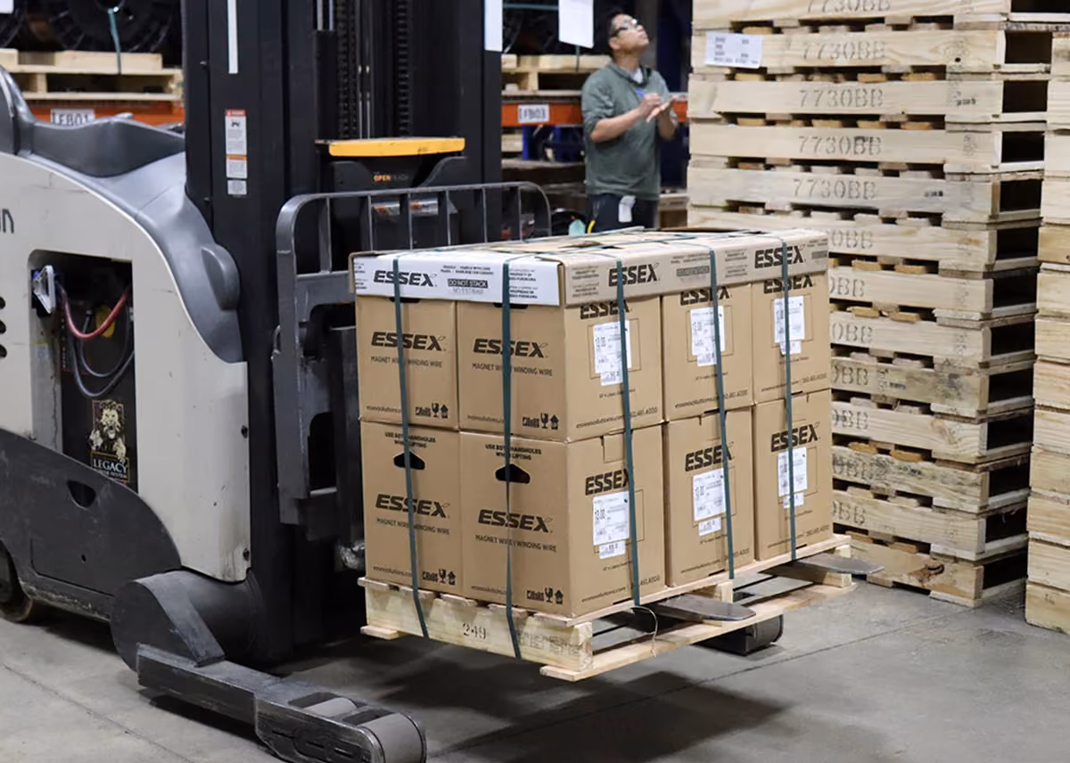 Forklift carrying pallet stacked with boxes labeled ESSEX magnet wire in warehouse.