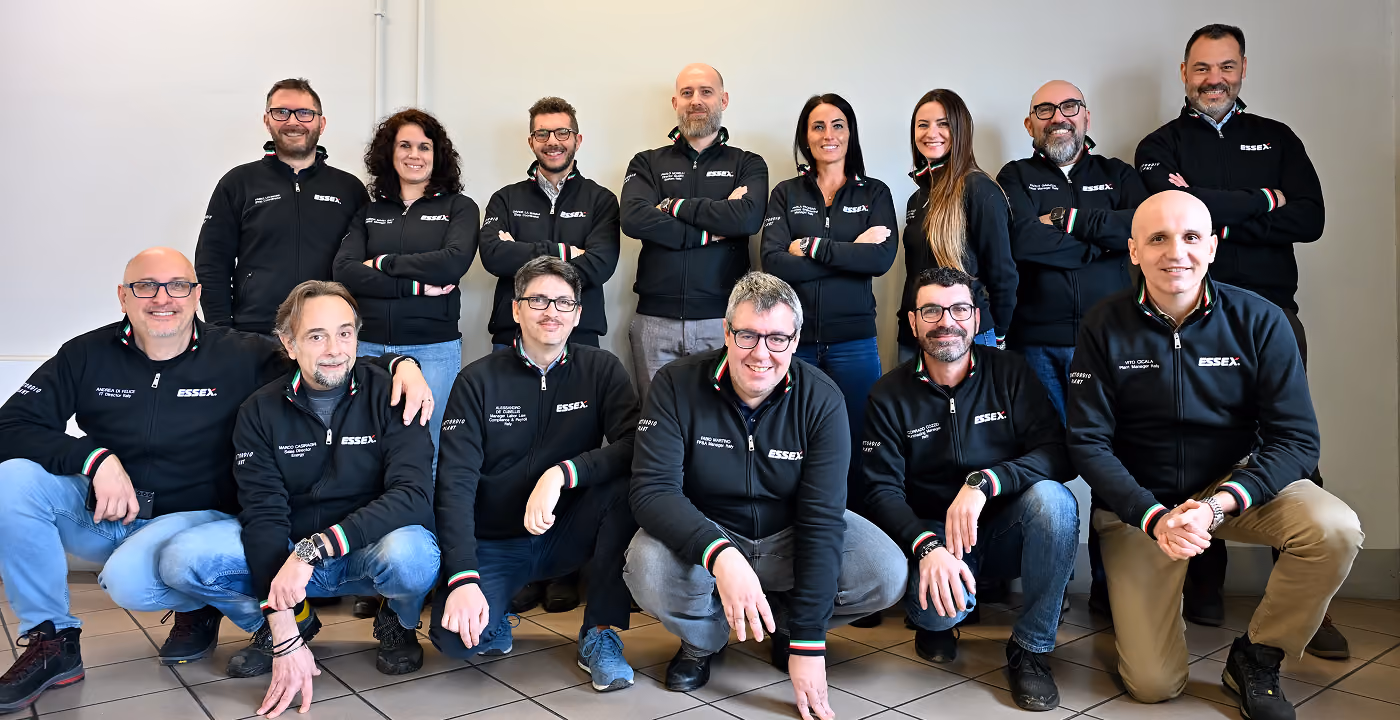 Group portrait of 14 adults in black Essex jackets smiling indoors against a plain wall.