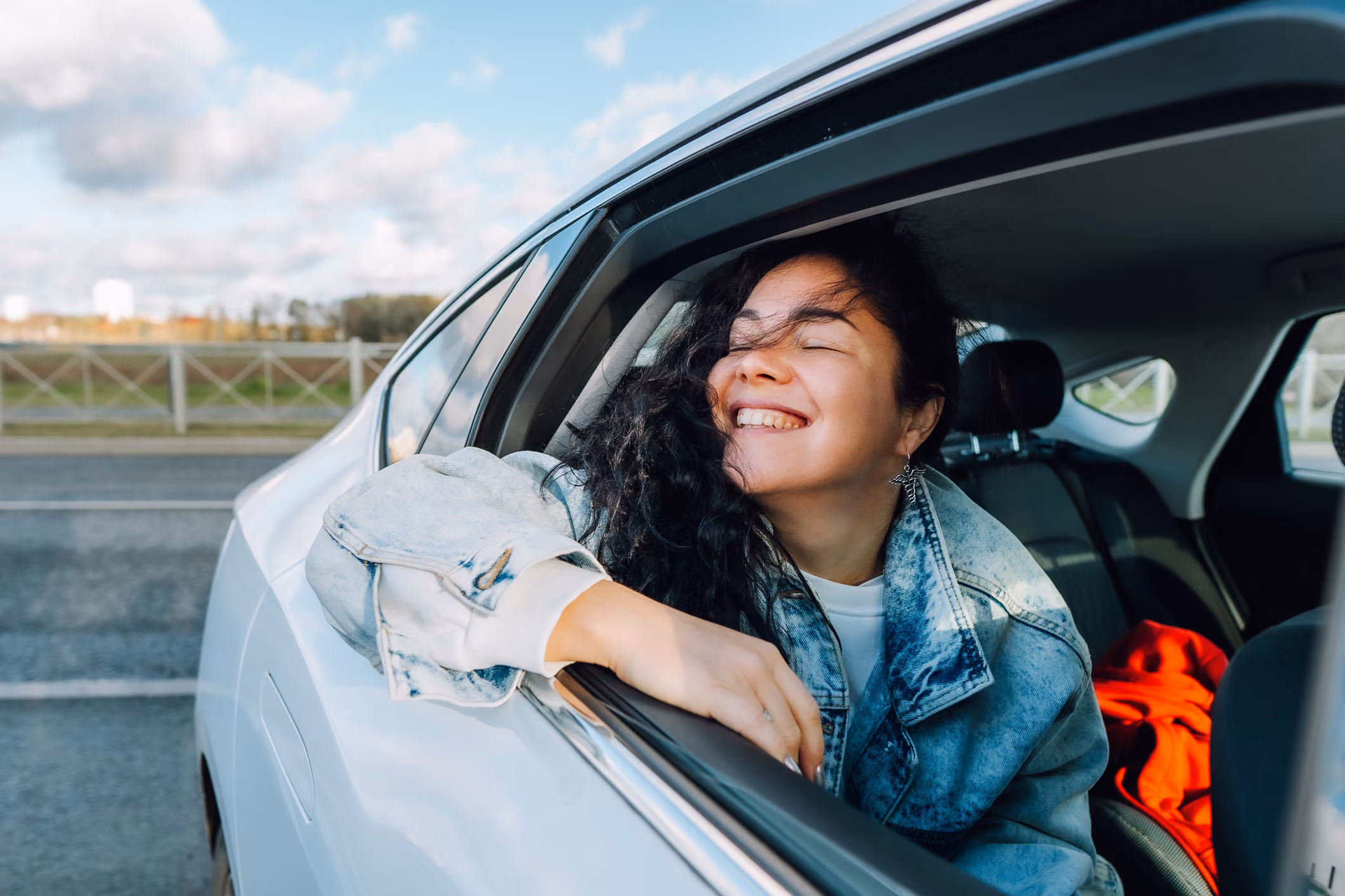 Smiling woman with curly hair leans out of a car window wearing a denim jacket.