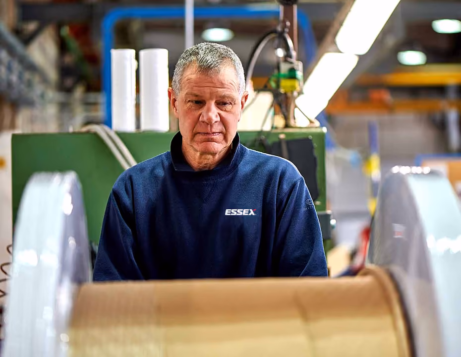 Man in navy ESSEX sweatshirt looking at large spool in an industrial setting.