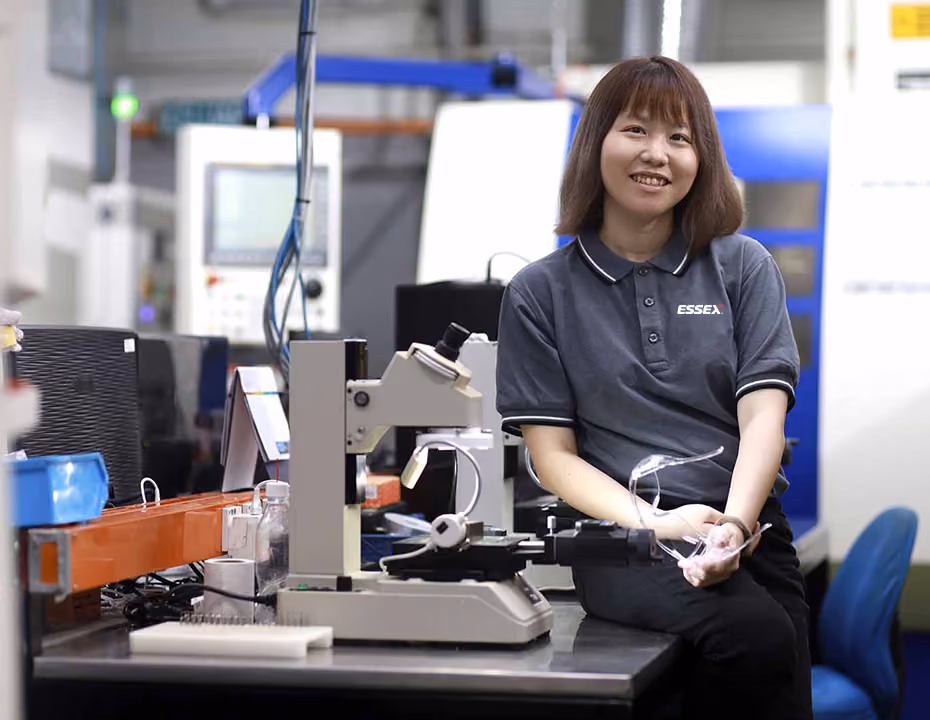 Smiling woman in dark shirt with Essex logo sitting by laboratory equipment in a scientific workspace.