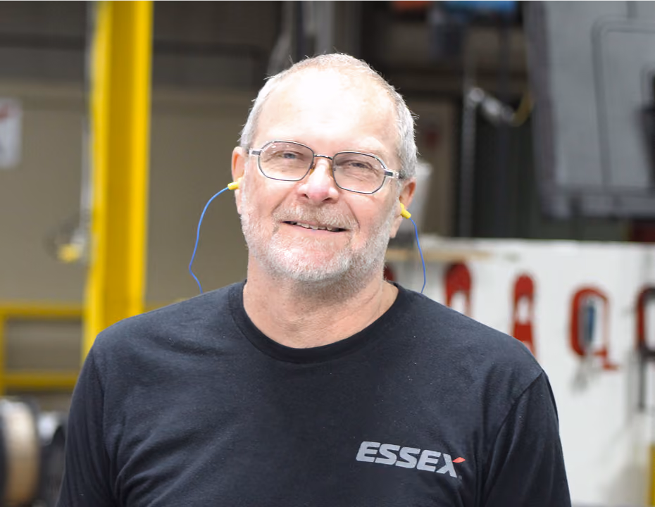 Smiling older man with glasses wearing a black Essex shirt and yellow earplugs in a factory setting.