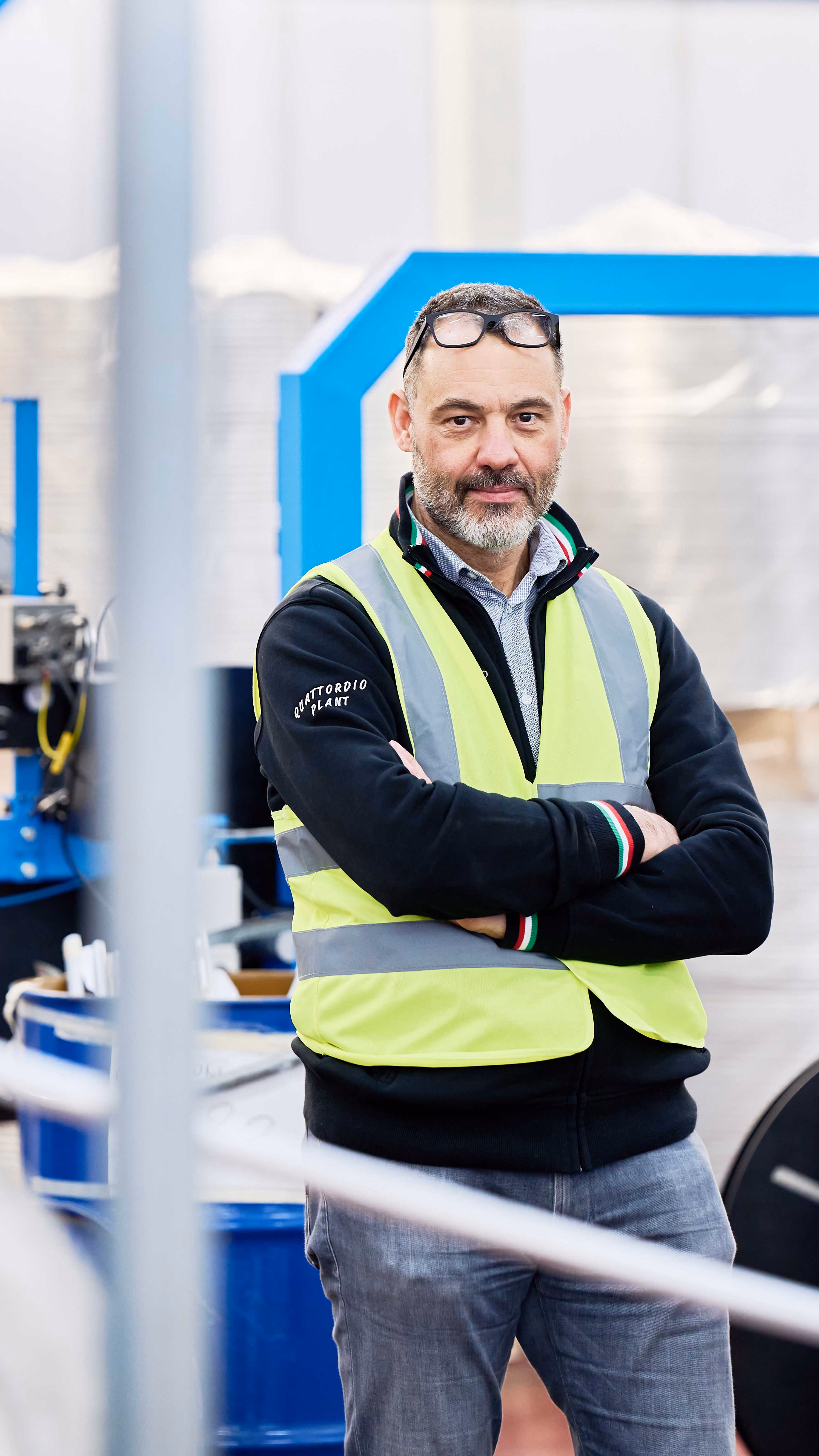 Man with beard wearing high-visibility vest and glasses on head standing with arms crossed in a factory.