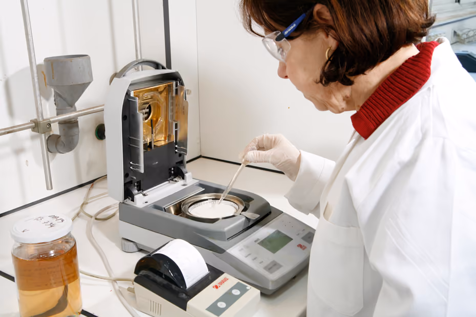 Scientist in lab coat and gloves using a moisture analyzer with a sample dish in a laboratory.