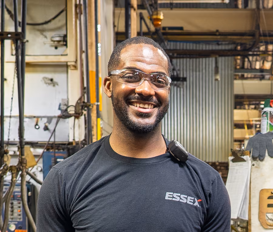 Smiling man wearing safety glasses and a black Essex t-shirt in an industrial workplace.
