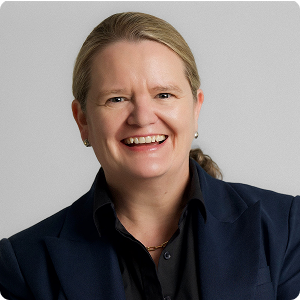 Smiling woman with light hair pulled back wearing a dark blazer and black shirt against gray background.