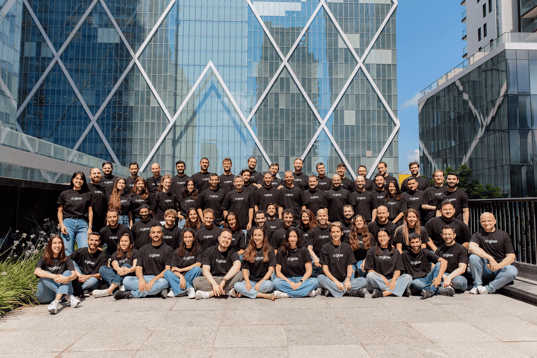 Large group of people wearing Glow black T-shirts posing in front of modern glass buildings outdoors.
