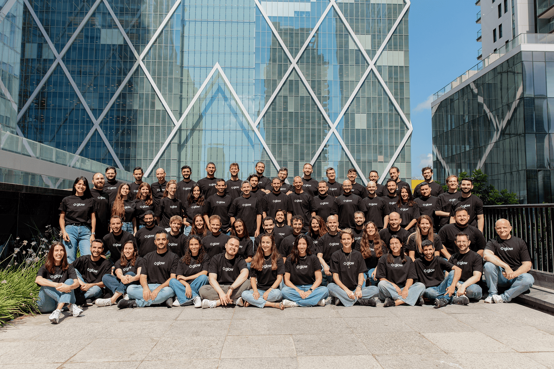 Large group of people wearing Glow black T-shirts posing in front of modern glass buildings outdoors.