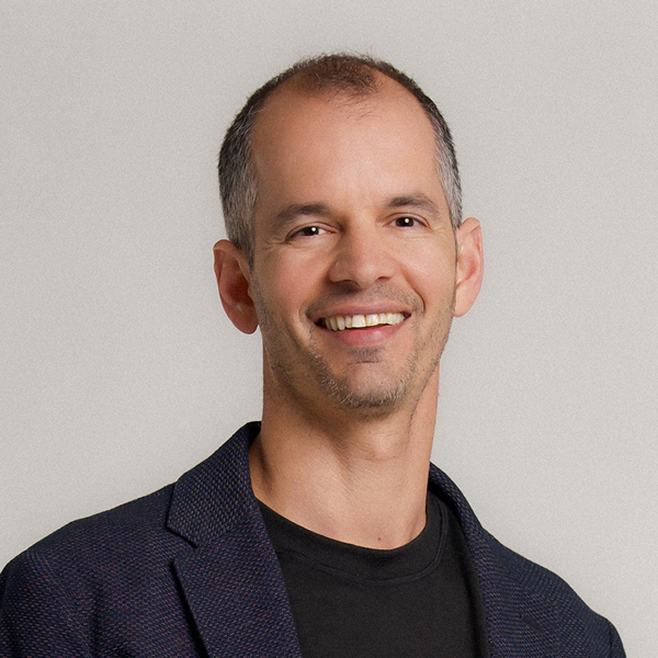 Smiling middle-aged man with short hair wearing a dark blazer and black shirt against a plain background.