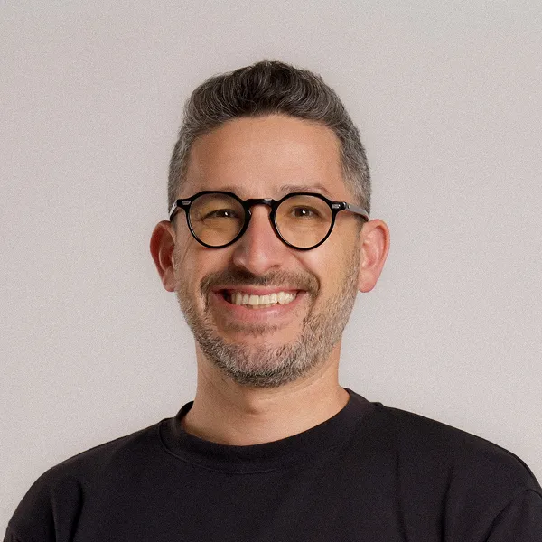 Smiling man with short graying hair, beard, black round glasses, and a black shirt on a plain background.