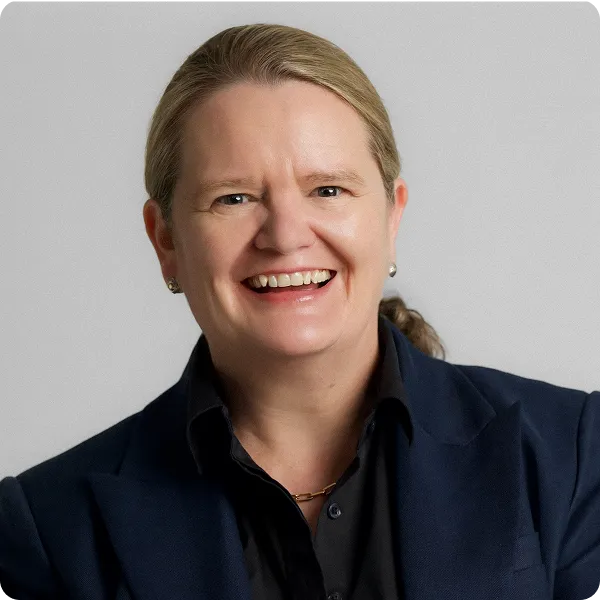 Smiling woman with light hair in a dark blazer and shirt against a plain gray background.