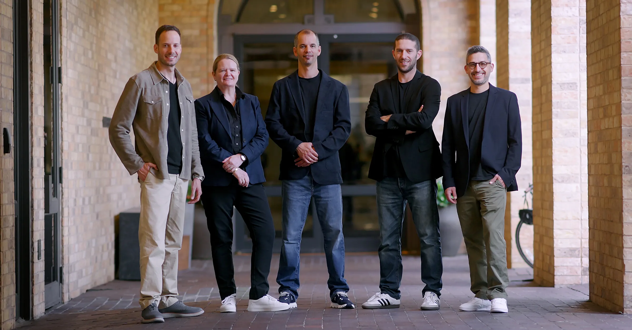 Five people standing in a row under an archway, dressed in casual blazers and sneakers, smiling.