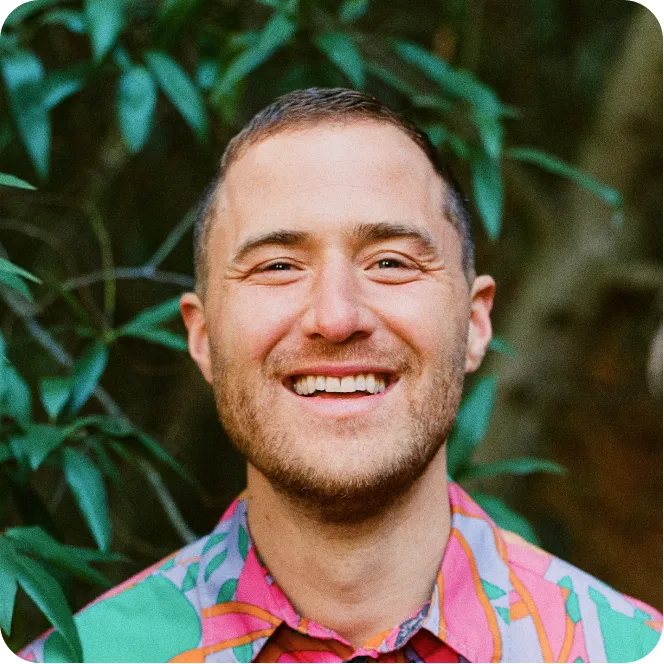 Smiling man with short hair and beard wearing a colorful patterned shirt with greenery in the background.