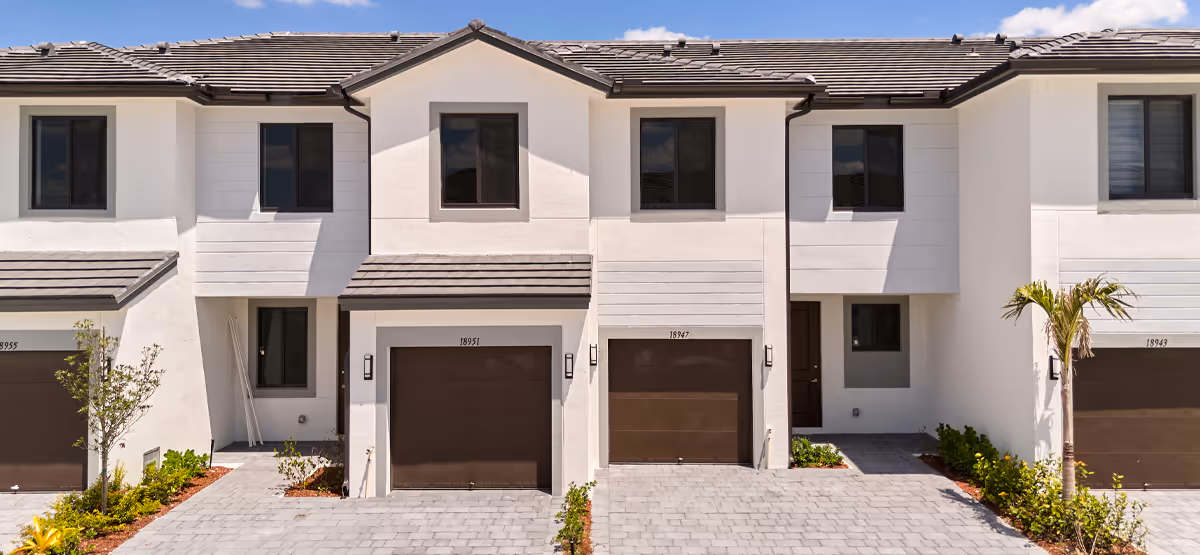 Row of modern two-story townhouses with white walls, dark garage doors, and tiled roofs under a blue sky.