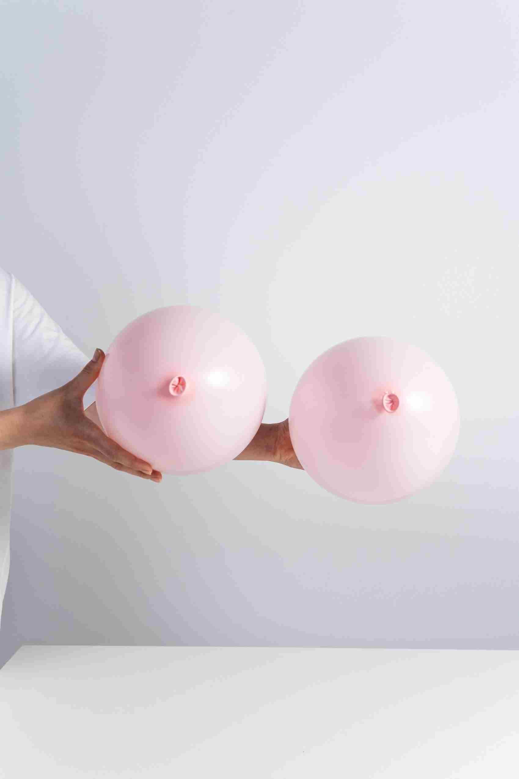 A woman holds two balloons that are staged to represent breasts on a white background