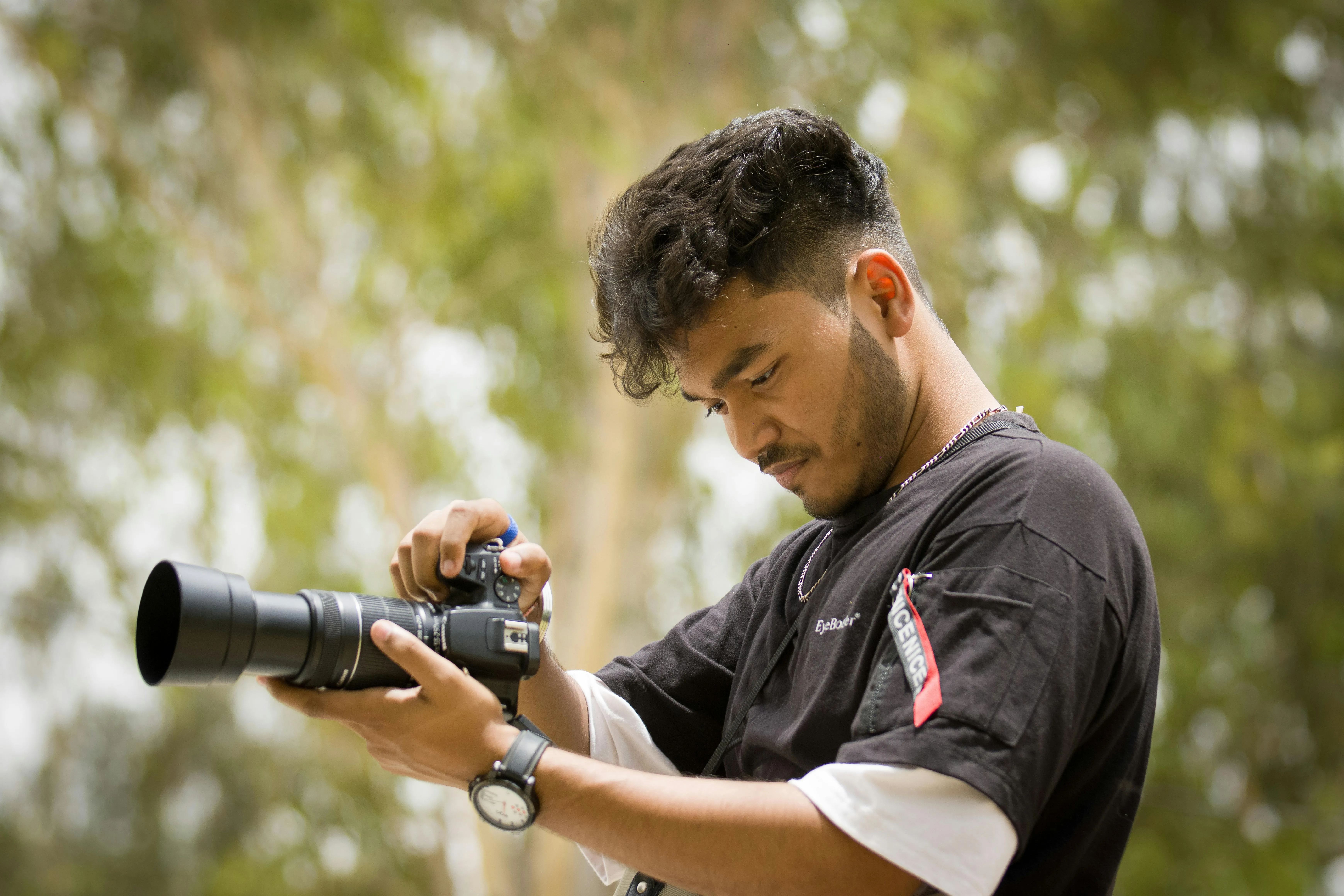 Young man outdoors adjusting settings on a DSLR camera with a long lens, blurred green background.