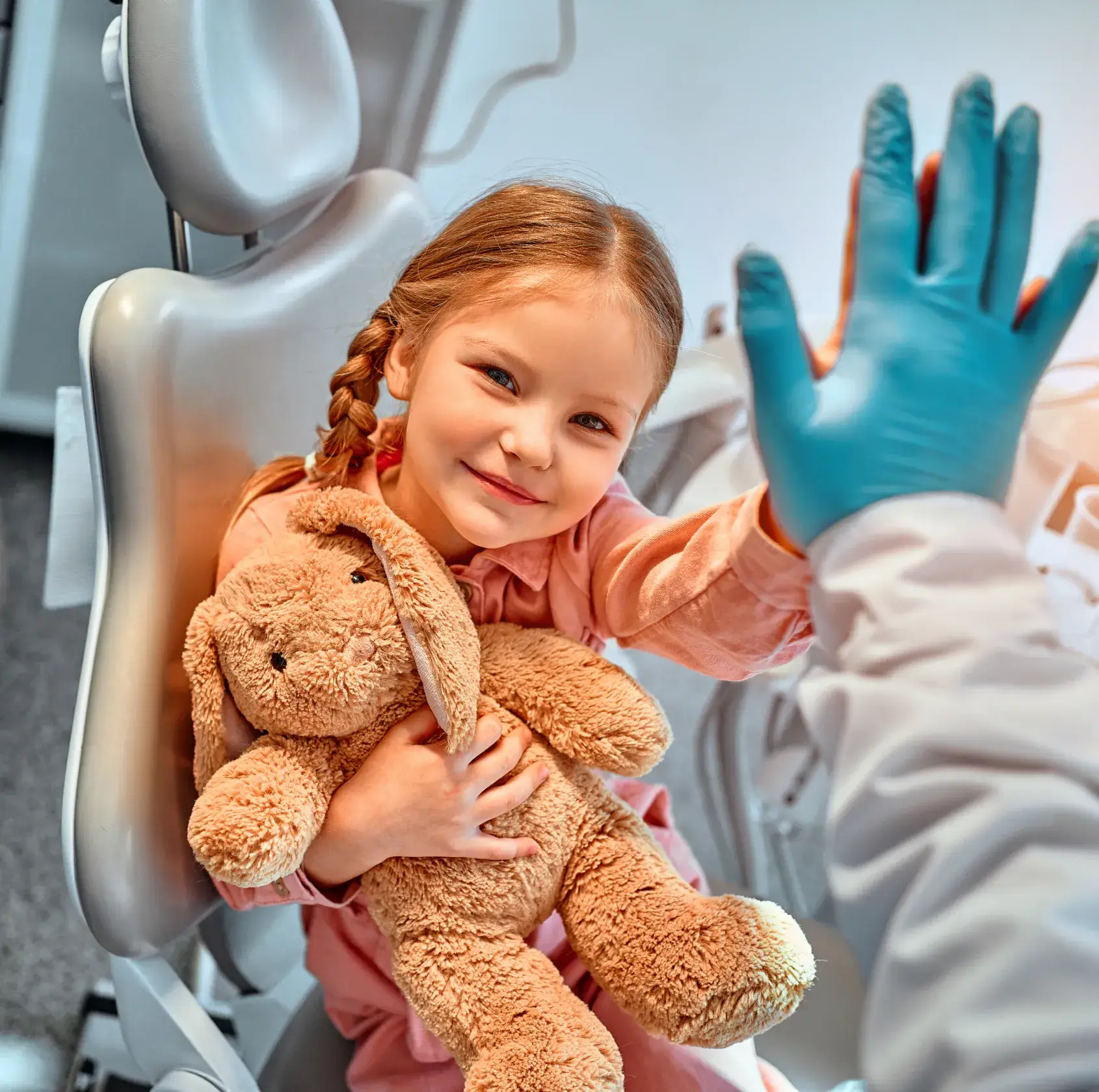 A little girl sitting in a dental chair holding a teddy bear.