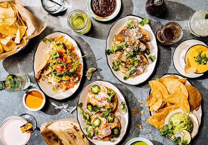 Mexican food on various plates including chips, tacos, salsa, guacamole and drinks
