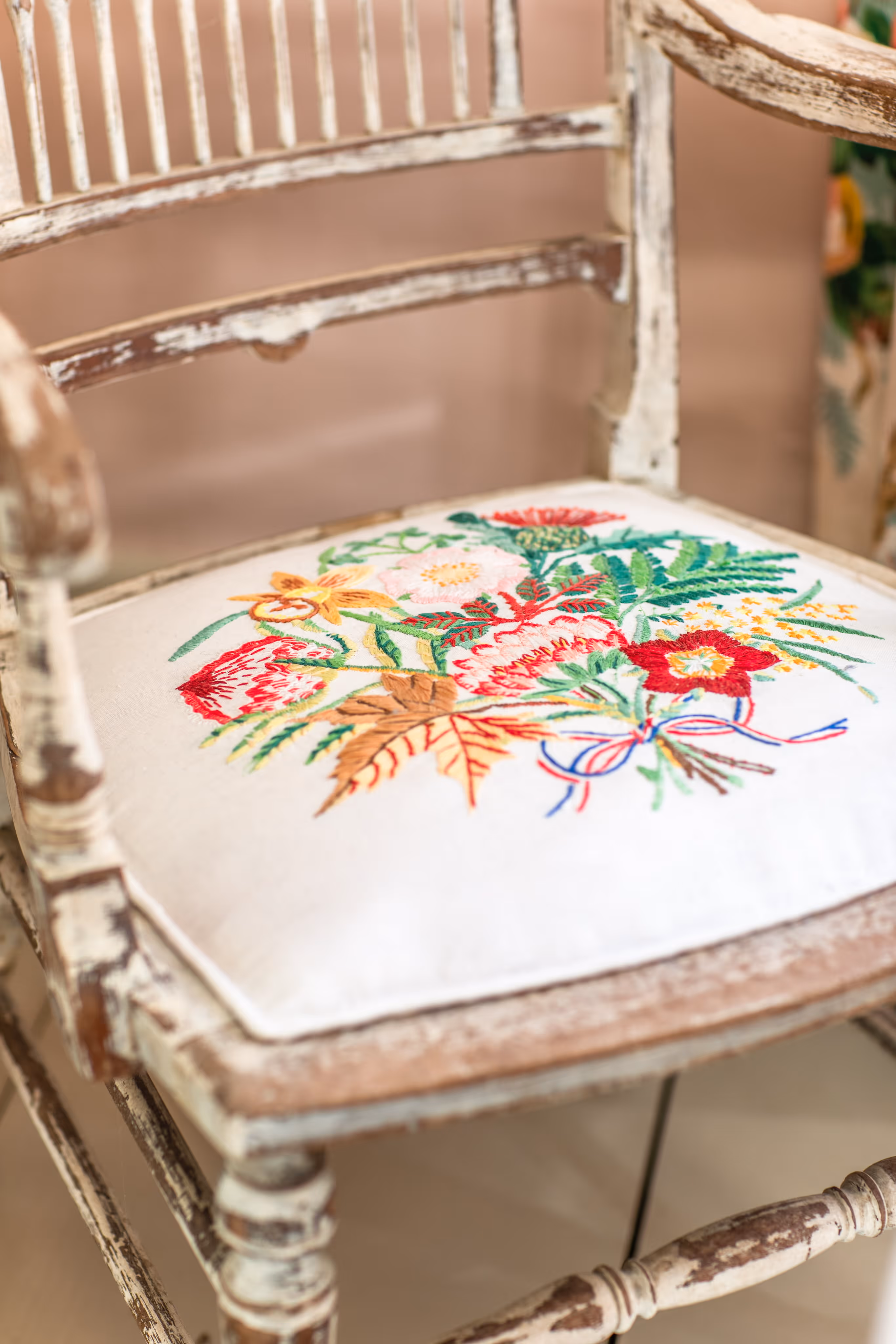 Close-up of a rustic wooden chair with white distressed paint and a white seat cushion embroidered with colorful flowers and leaves.