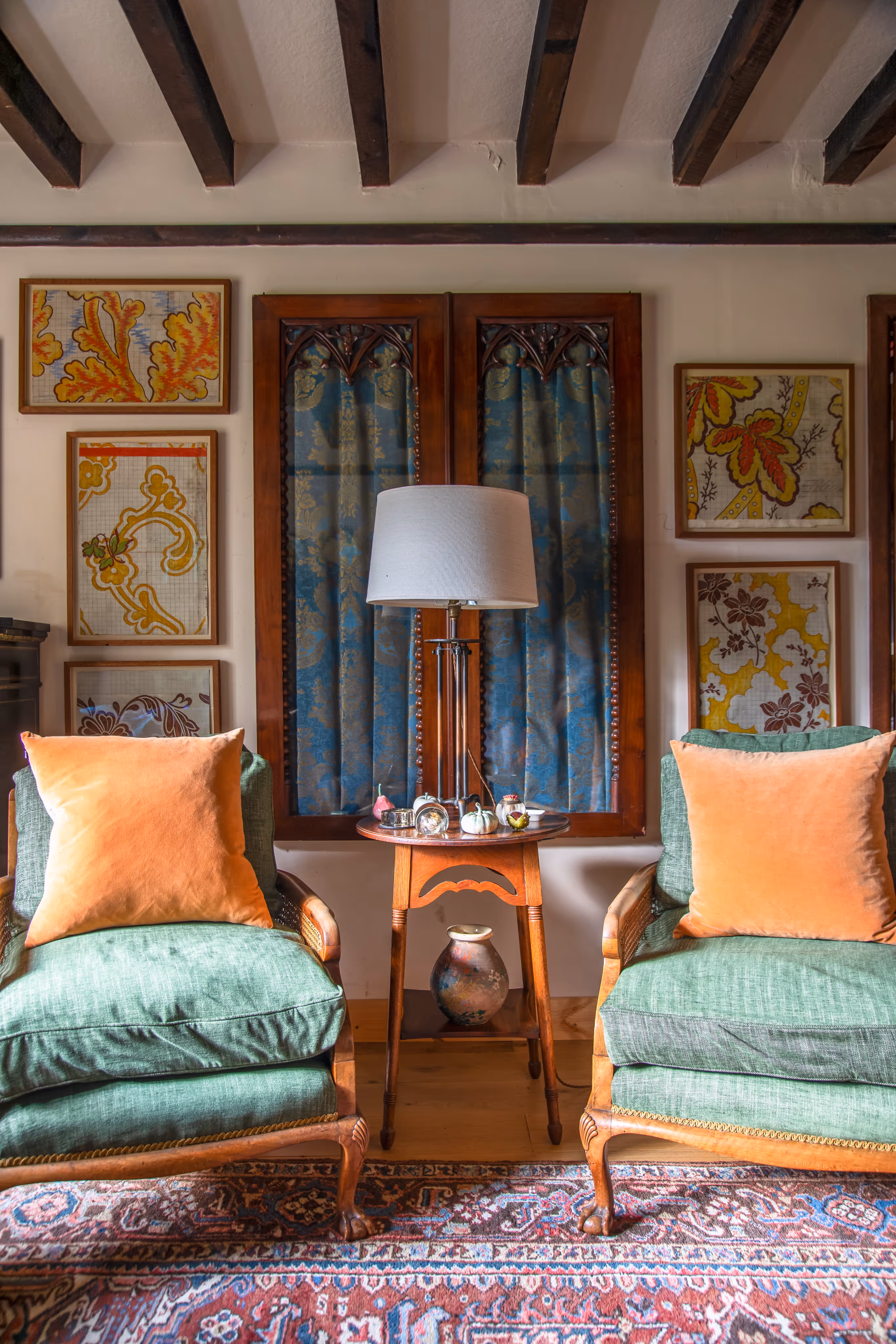 Cozy sitting area with two green cushioned wooden armchairs with orange pillows, a small wooden side table with a white lampshade and decorative items, framed textile art on the walls, and exposed wooden ceiling beams.