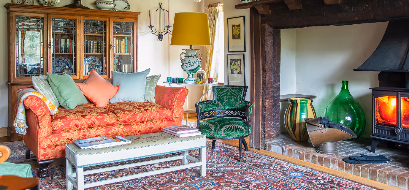Cozy living room with a patterned red sofa, green armchair, glass-front cabinet, fireplace with fire burning, and decorative glass vases.