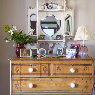 Wooden dresser with white knobs topped with framed photos, a vase of flowers, a glass lamp with a pleated shade, and a white decorative mirror above.