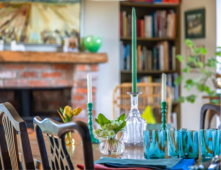 Dining table set with blue glasses, floral centerpiece, and green candles in glass holders, with a brick fireplace and bookshelf in the background.