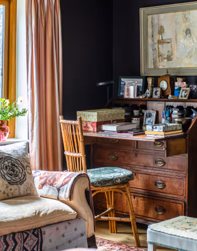 Cozy room corner with a wooden desk cluttered with books, photos, and office supplies, a vintage chair, and a cushioned sofa near a window with pink curtains.