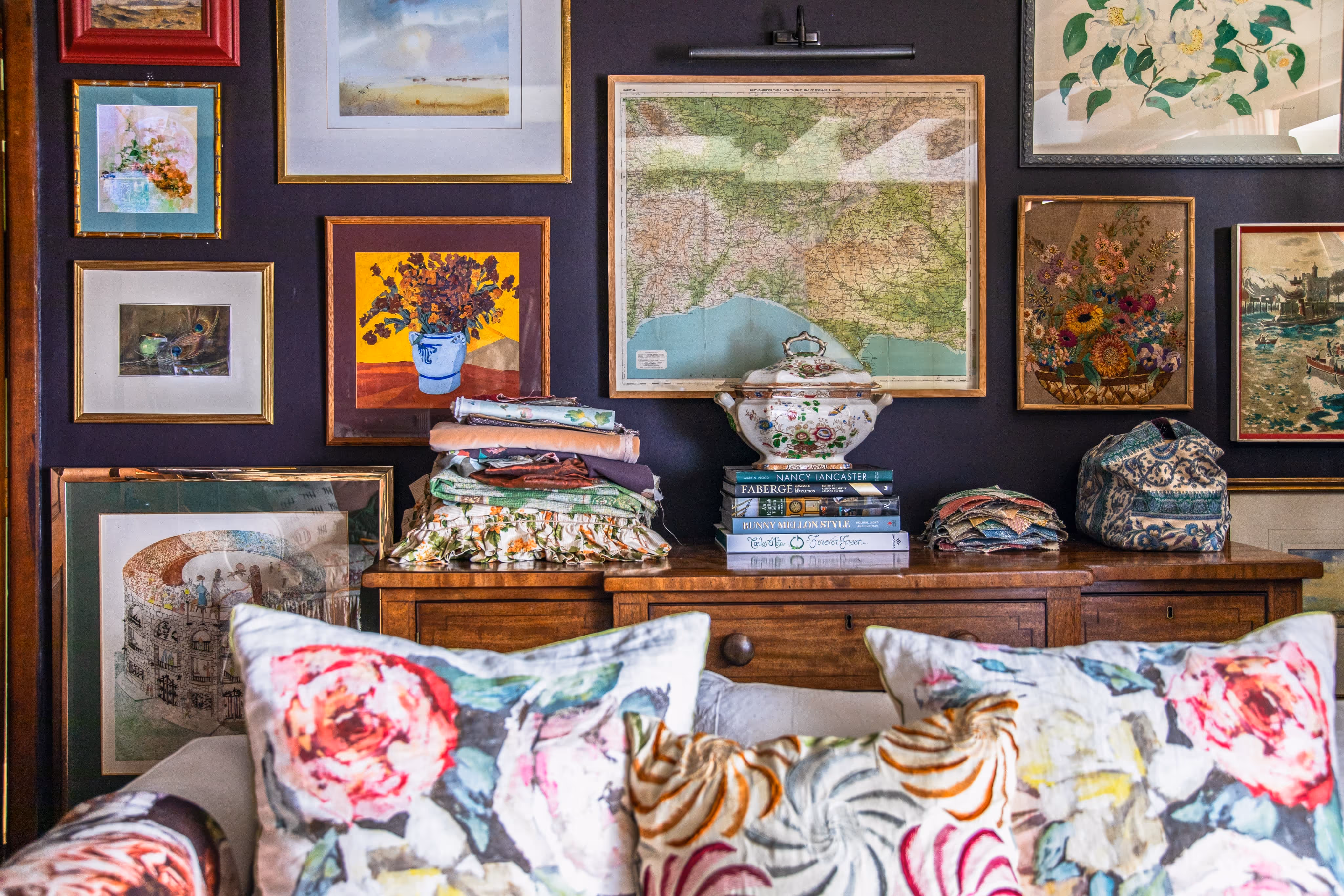 Wall decorated with various framed paintings and a vintage map above wooden furniture holding folded fabrics, books, and a floral porcelain tureen, with floral-patterned cushions in the foreground.