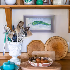 Kitchen shelf displaying wooden cutting boards, cooking utensils in a white holder, decorative ceramic bowls, and a painting of a green chili pepper on the wall.