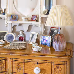 Wooden dresser with decorative carvings holding a pink glass lamp with a pleated shade, framed photos, a silver dish with red accents, and fine china teacups.
