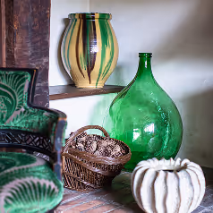 Decorative green and beige ceramic vases and a basket with pine cones arranged on a rustic floor and shelf.