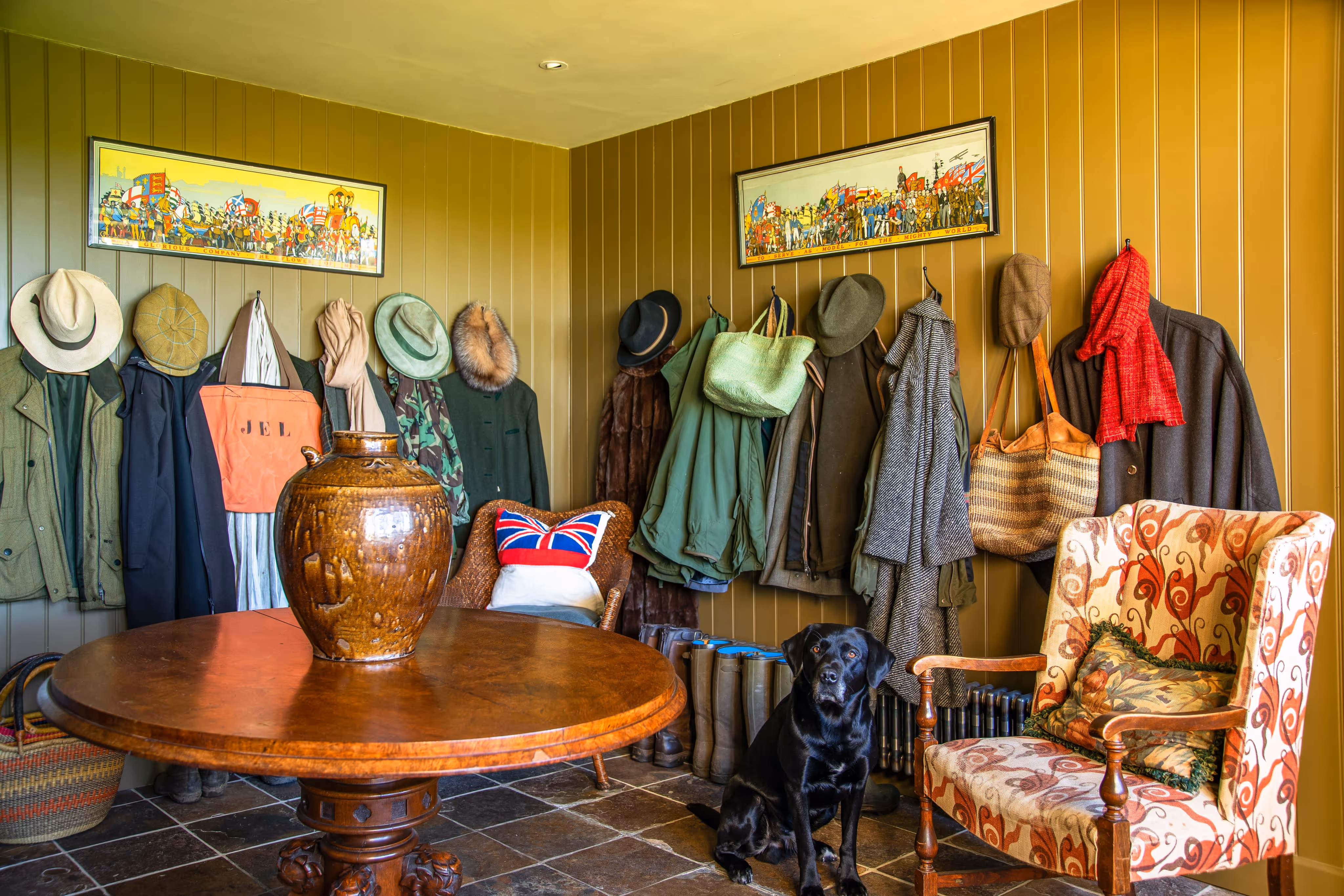 Cozy room corner with wooden round table, large brown vase, coats and hats hanging on walls, patterned armchair, and black Labrador sitting on tiled floor.