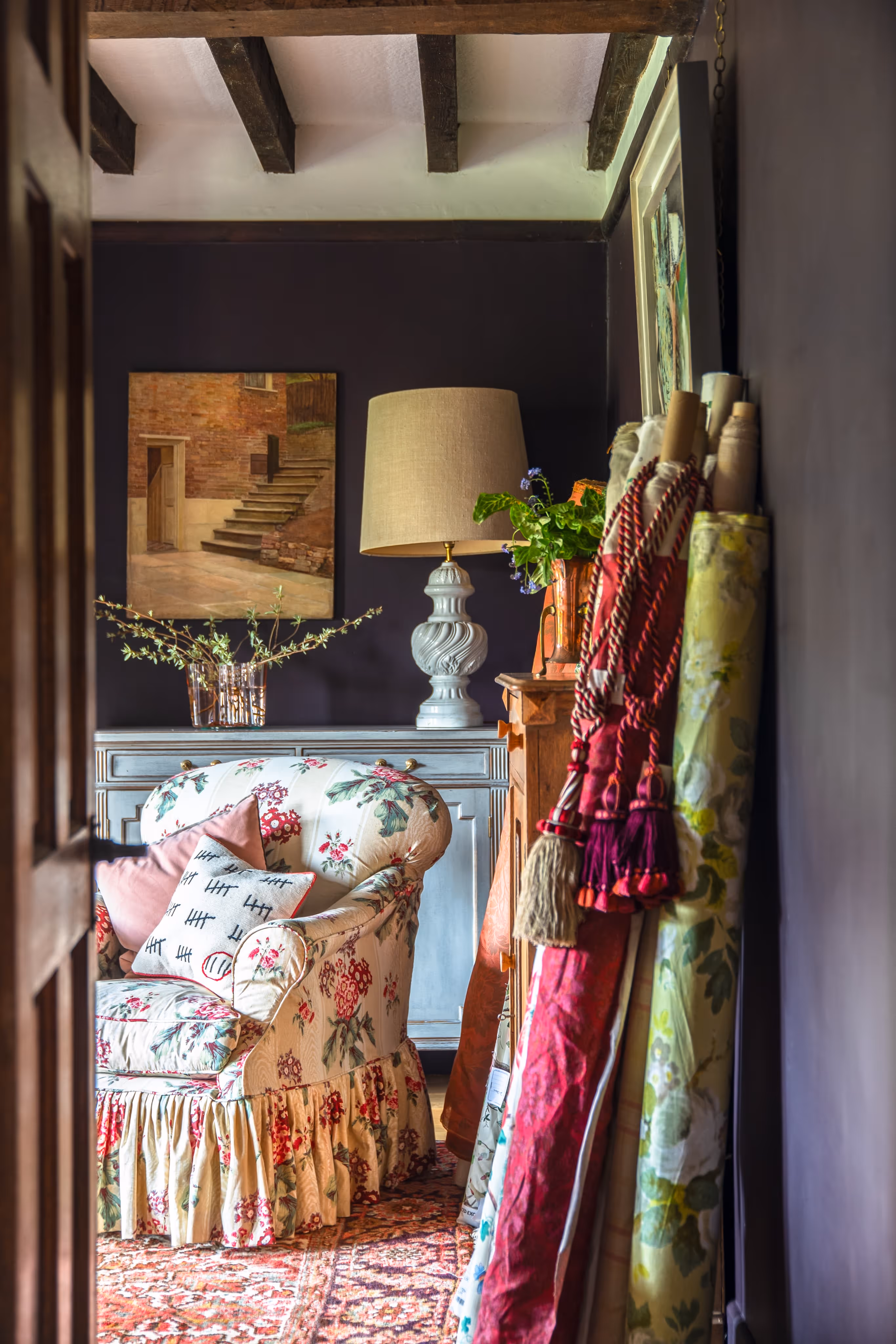 Cozy room with floral armchair, patterned rug, wooden beams, cabinet with lamp and plants, and rolls of fabric with decorative tassels.