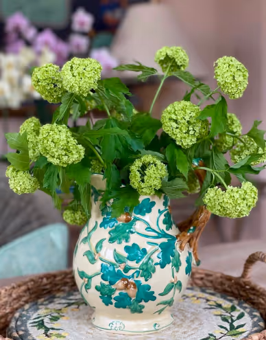 Decorative ceramic vase with green floral patterns holding a bouquet of green hydrangea flowers on a round woven tray.