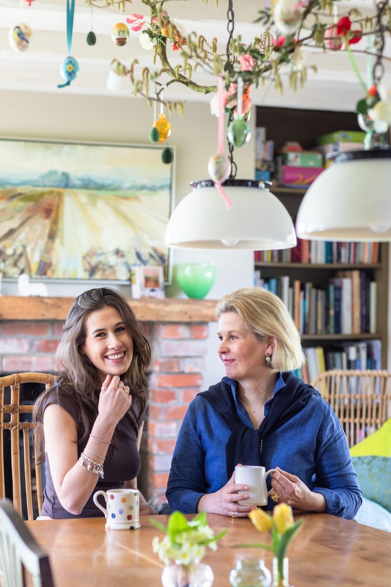 Two women sitting at a wooden table in a cozy room with hanging decorations, smiling and holding mugs.