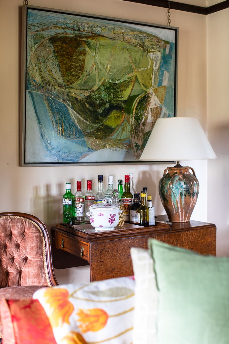 Interior corner with a wooden side table holding assorted liquor bottles, a floral ceramic bowl, and a large ceramic lamp, beneath a framed abstract painting.