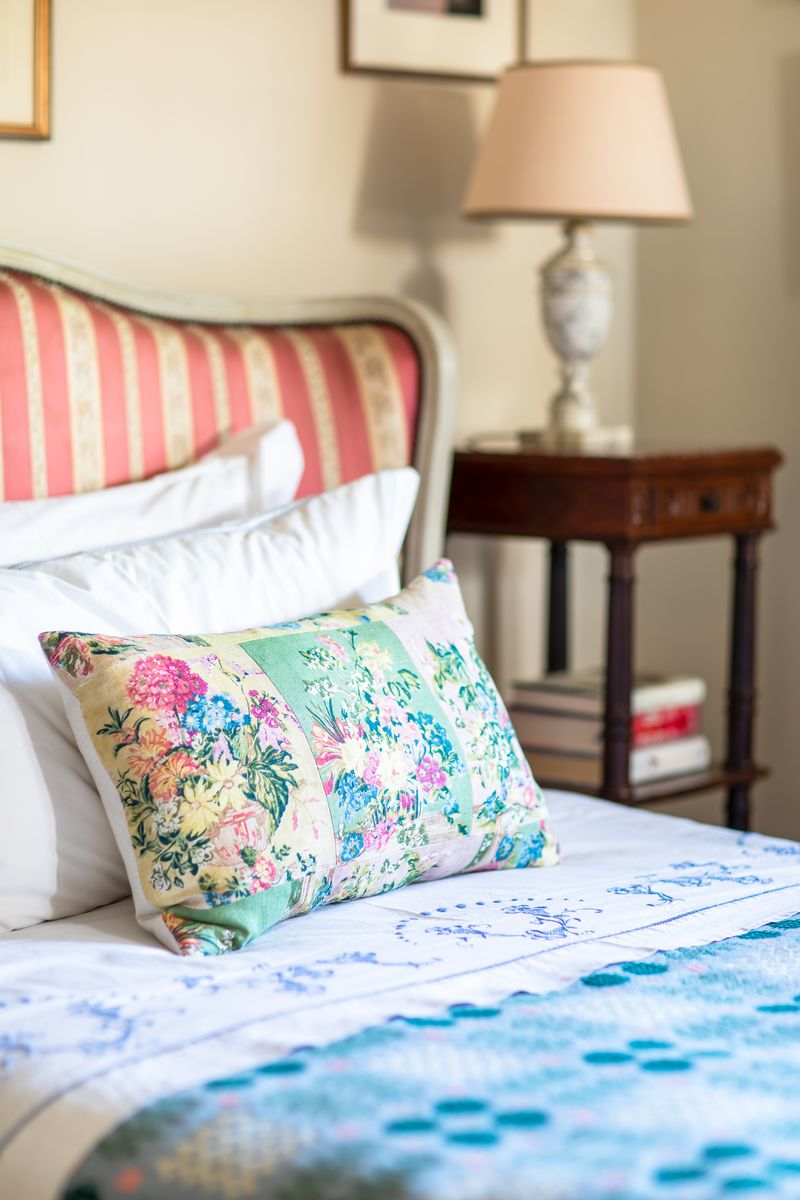 Bed with a floral patterned pillow, white sheets, a striped headboard, and a wooden nightstand with a lamp and books.