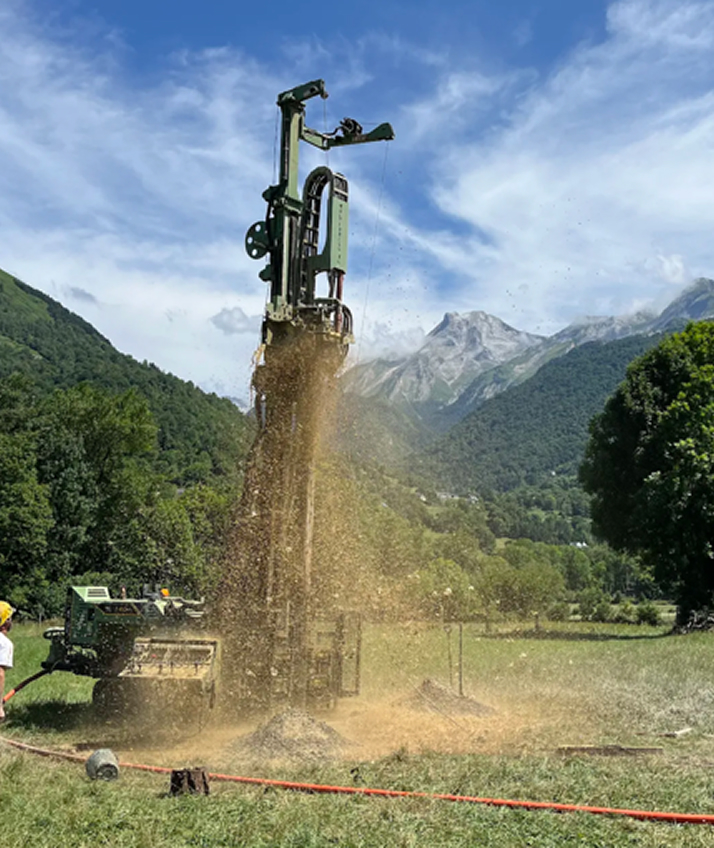 Drilling machine ejecting soil at a grassy mountain site under a partly cloudy sky.