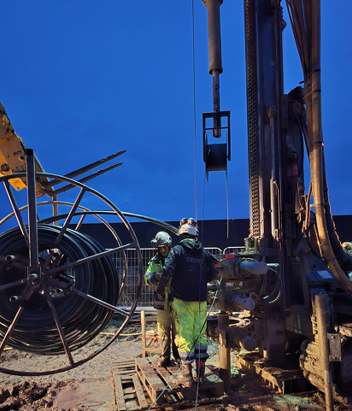 Two workers in helmets operating drilling machinery next to large cable reel during twilight.
